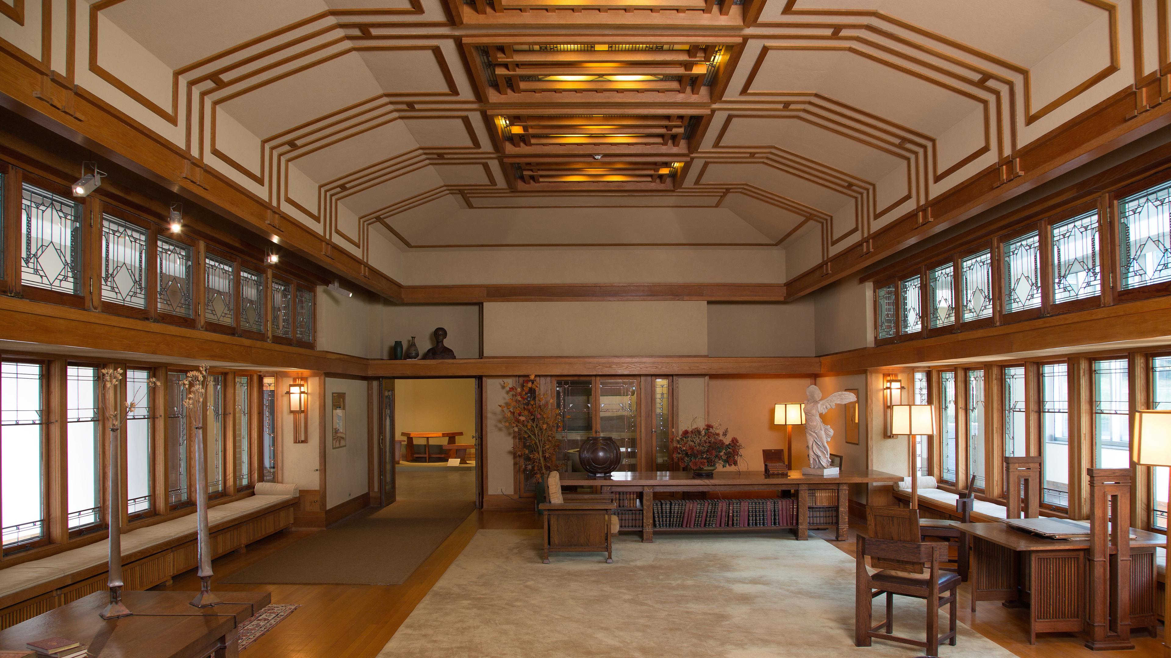 View of the Frank Lloyd Wright Room. An expansive living area, with windows on each side, and wood paneling paired with sturdy wood furniture.