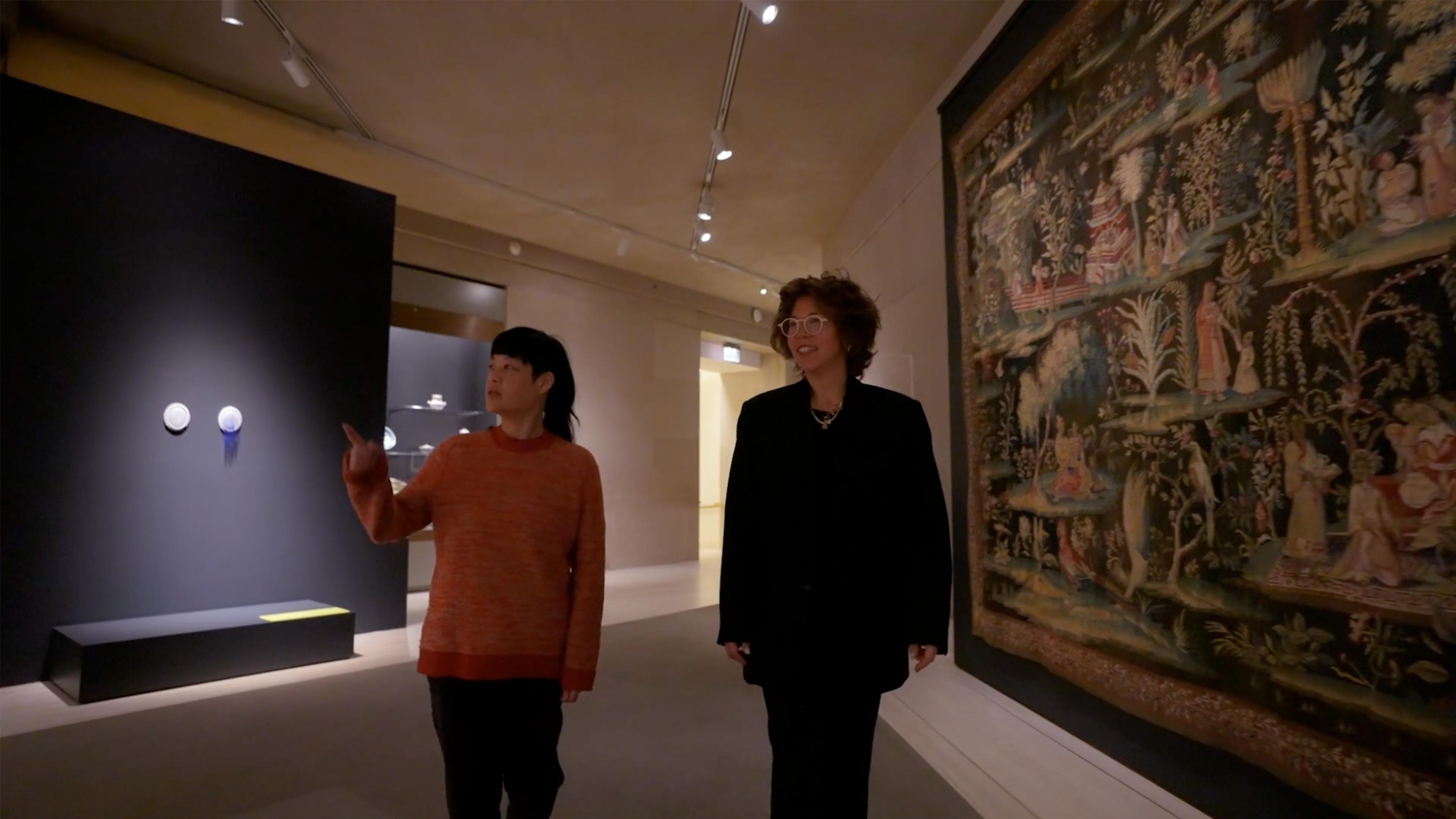 Two women one in red, and one in all black walking a dim lit gallery.