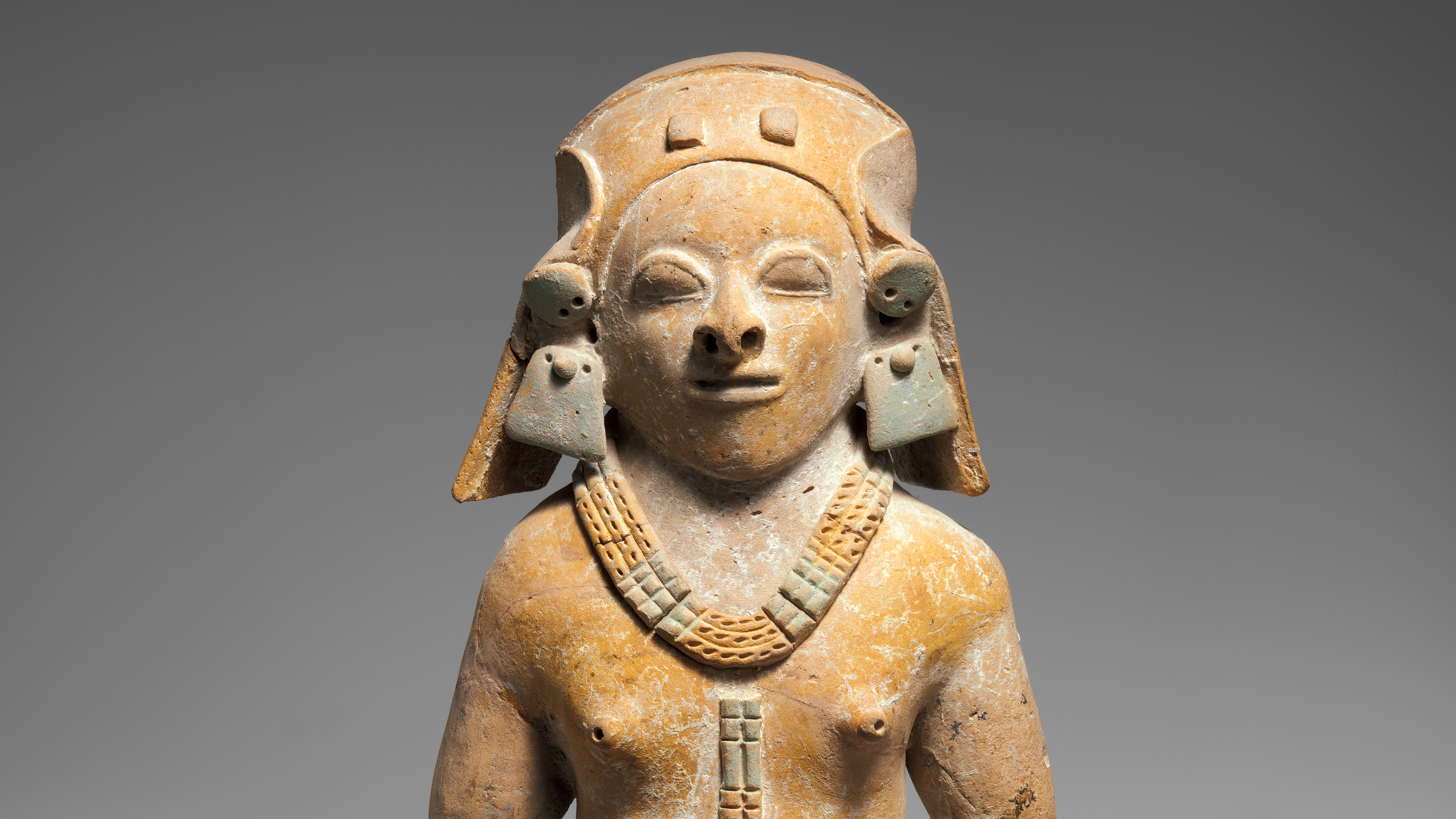 Bare-chested female ceramic figure wearing a multistrand necklace with a sizable pendant and a decorated head cover.