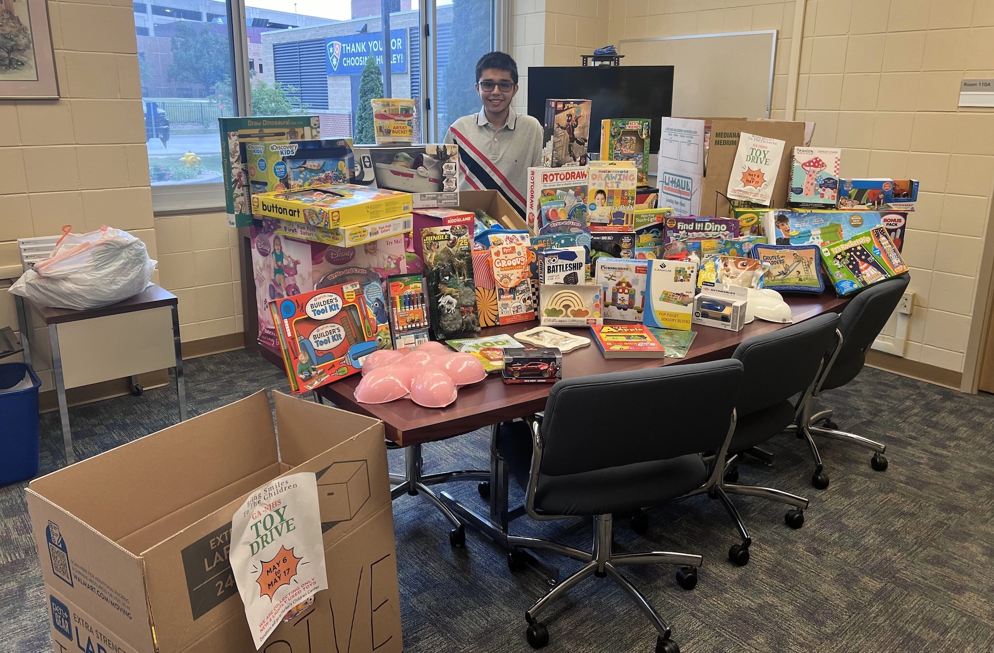 A community member stands behind a table filled with a variety of toys and games for a toy drive, promoting volunteerism and community support. A large cardboard box labeled for toy collection sits on the floor, highlighting the initiative's goal to serve children in need.