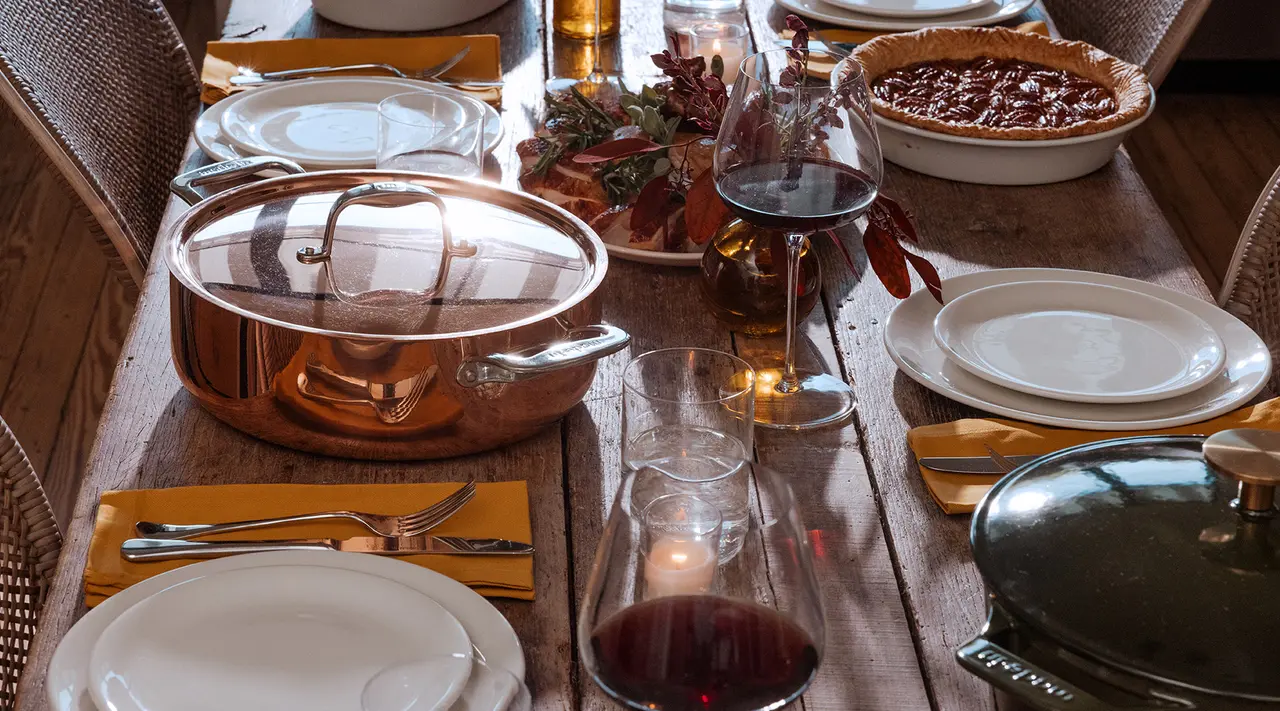 A cozy dinner setting with copper pots, wine glasses, and a pie on a rustic wooden table illuminated by soft, warm light.