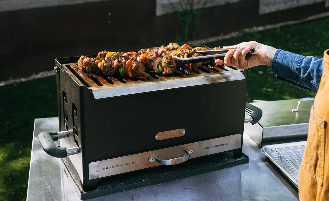 A person uses tongs to turn skewers of meat and vegetables on a grill in an outdoor setting.