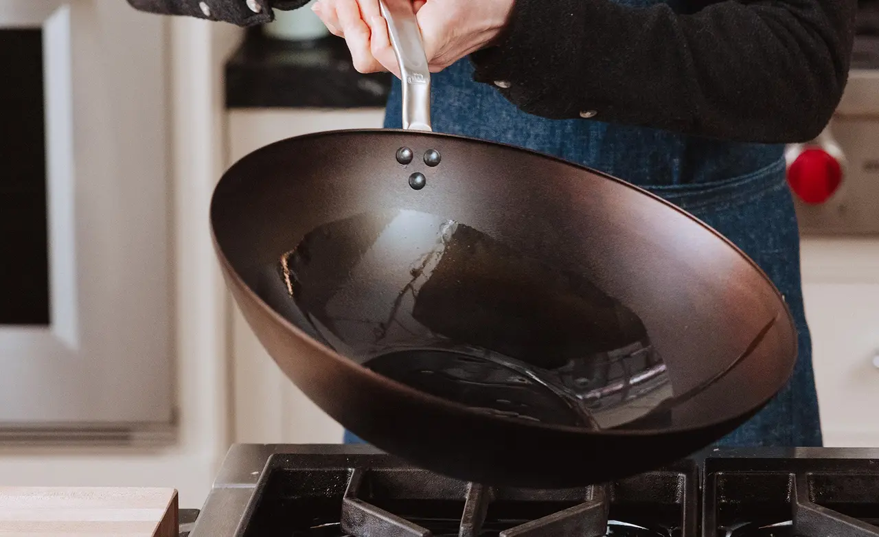 A person is holding a dark, shiny wok over a gas stovetop, preparing to cook.