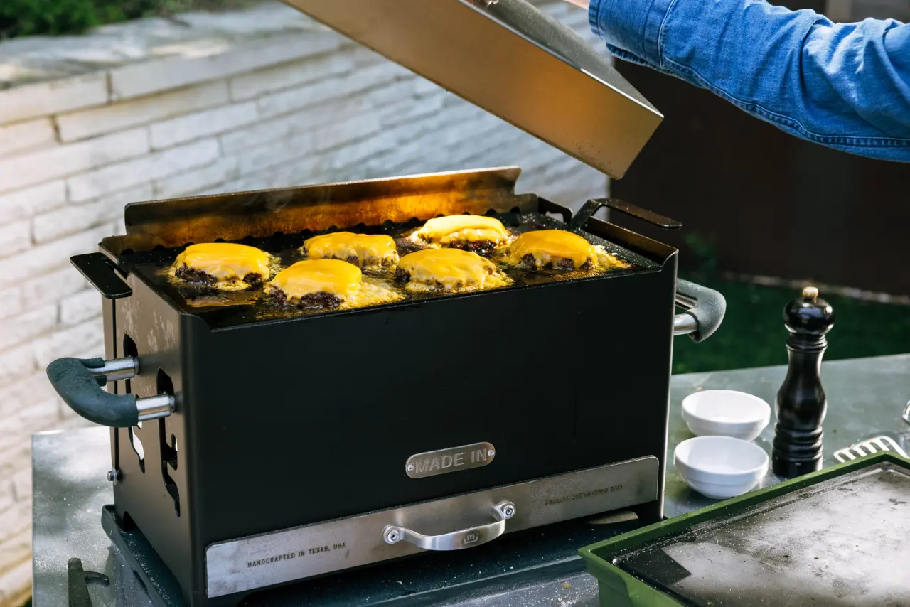 Cheeseburgers are cooking on a black griddle with a lid being lifted to check on the food.