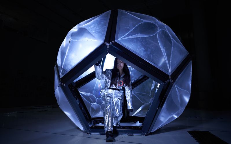 A person wearing a reflective silver suit steps out of a glowing geodesic pod-like structure in a dark space. The faceted dome is made of translucent panels with organic patterns illuminated from within, creating a futuristic and immersive atmosphere.