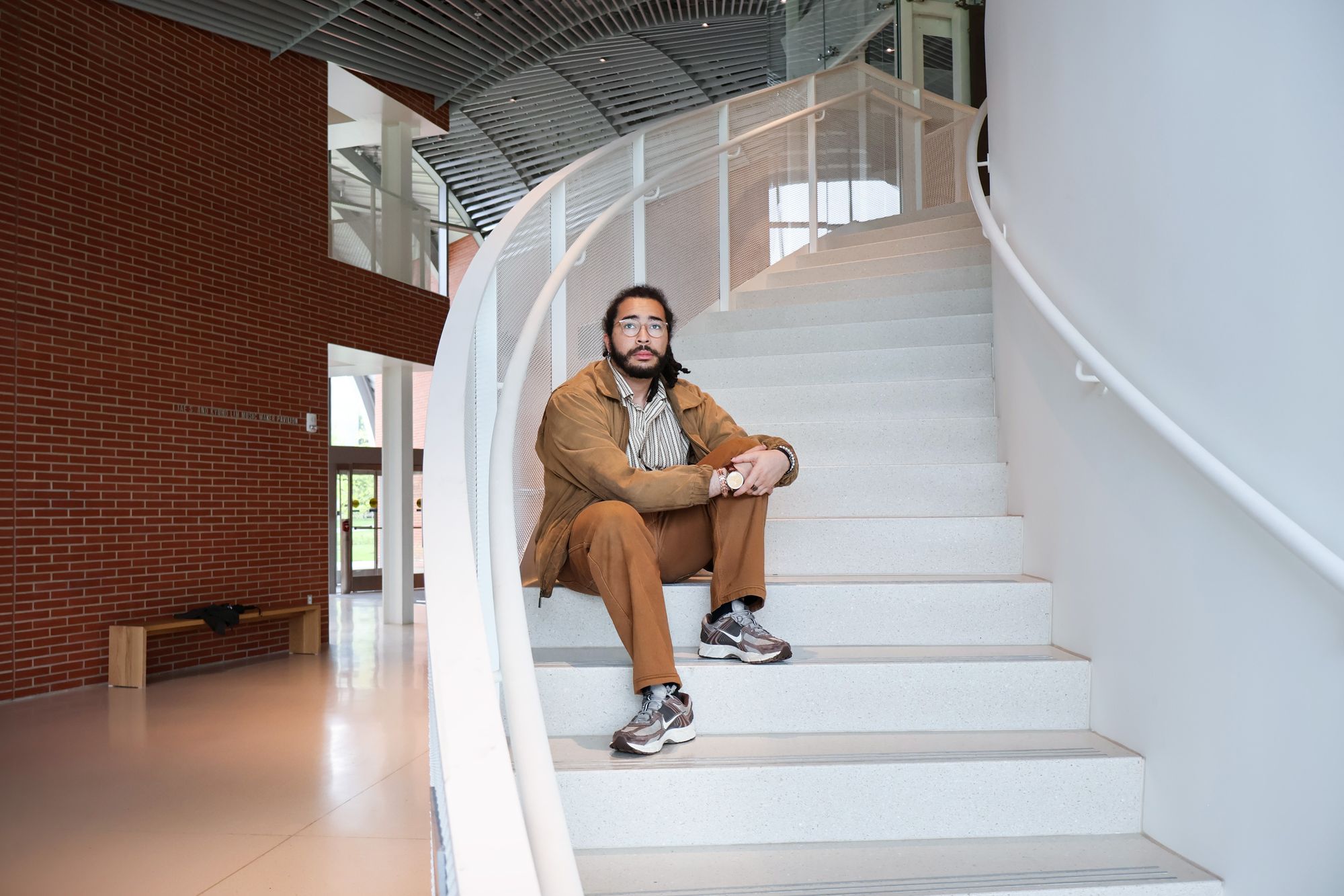 A man with medium-length hair and a beard sits on a curved indoor staircase. He is wearing glasses, a brown jacket, brown pants, and sneakers. The setting appears to be a modern building with red brick walls, large windows, and a light-colored floor.
